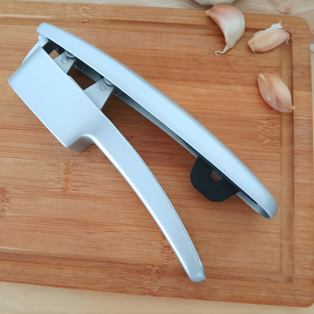 Knoblauchpresse in Silber auf einem Holzbrett, danebenliegend Knoblauchzehen.
